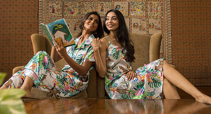 Two women sit on the floor in front of an armchair, wearing matching floral pajamas. One woman reads a book while they both smile at the camera. A patterned wall hanging decorates the background.