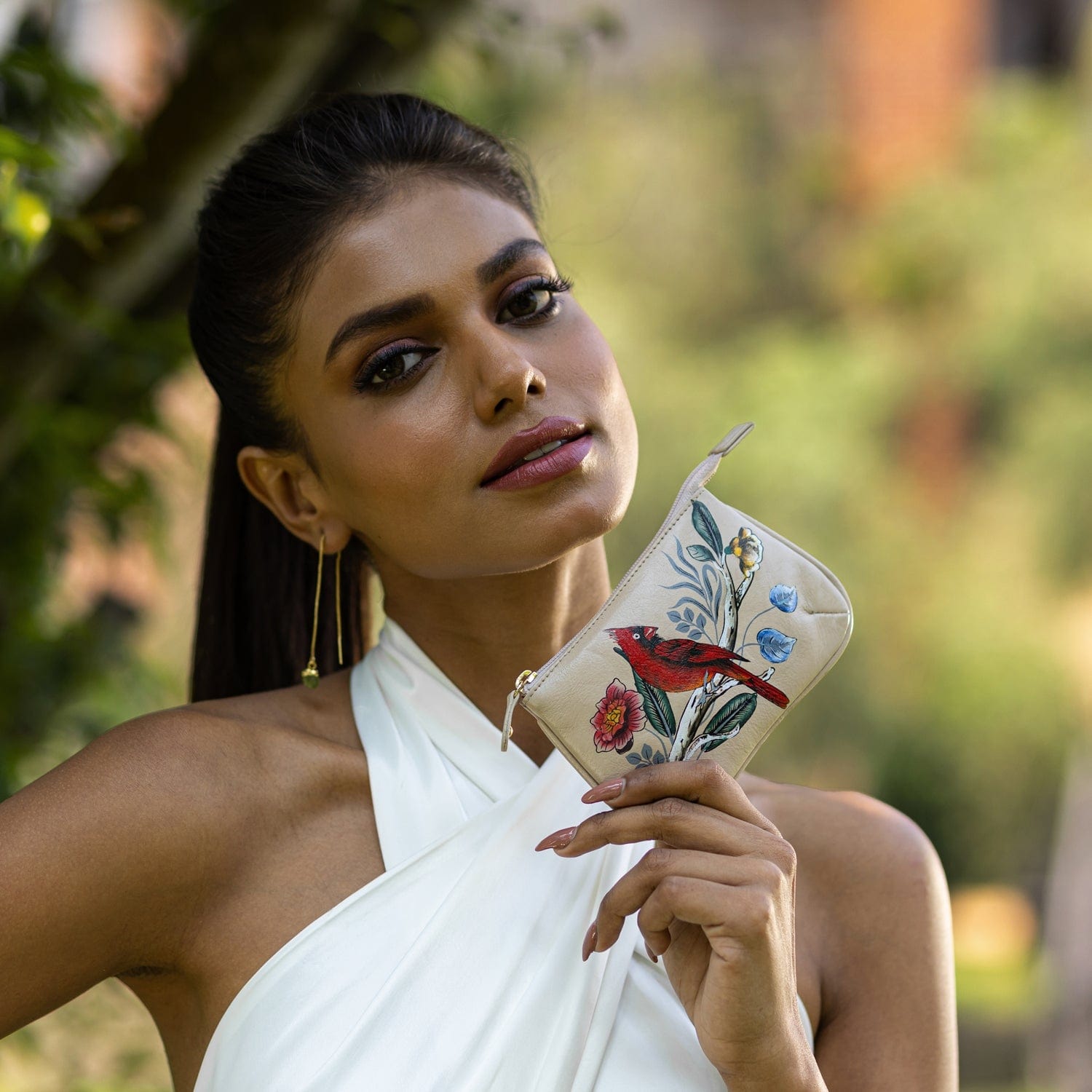 A woman in a white dress holding an Anuschka Medium Zip Pouch - 1107 with a bird design outdoors.