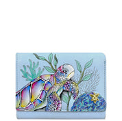 The Anuschka RFID Blocking Small Flap French Wallet - 1138 is a light blue genuine leather wallet adorned with hand-painted artwork of a colorful sea turtle and ocean plants, featuring RFID protection and a small starfish decoration on the bottom right corner.