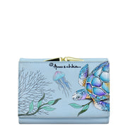 The Anuschka RFID Blocking Small Flap French Wallet - 1138 is a light blue wallet featuring hand-painted artwork of an ocean scene with a sea turtle, jellyfish, and seaweed. Crafted from genuine leather, it includes RFID protection for security and proudly displays the word "Anuschka.