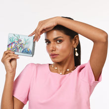 Load image into Gallery viewer, A woman in a pink top holds the Anuschka RFID Blocking Small Flap French Wallet - 1138, featuring a hand-painted fish and coral design, her left arm raised to touch her head. Made from genuine leather, the wallet offers RFID protection. She accessorizes with pearl earrings and a matching necklace.
