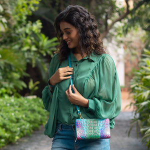A woman in a green blouse and jeans, carrying a colorful bag with an Anuschka Organizer Wallet Crossbody - 1149 inside, smiling while looking down.