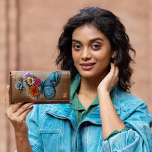 Load image into Gallery viewer, A person in a jean jacket holds up an Anuschka Triple Fold RFID Clutch Wallet - 1150, which features colorful, hand-painted artwork of a butterfly and flowers, while smiling at the camera.
