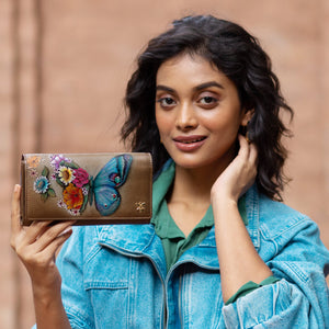 A person in a jean jacket holds up an Anuschka Triple Fold RFID Clutch Wallet - 1150, which features colorful, hand-painted artwork of a butterfly and flowers, while smiling at the camera.