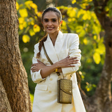 Load image into Gallery viewer, A woman in a white blazer and braided hair, holding an Anuschka Crossbody Phone Case - 1173, stands confidently in an outdoor setting with trees and yellow foliage in the background.
