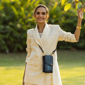 A woman in a cream suit smiling outdoors with a small blue Crossbody Phone Case - 1173 featuring an RFID card holder from Anuschka.