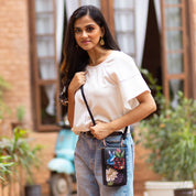 A woman stands outdoors holding an Anuschka Crossbody Phone Case - 1173 with floral designs and a crossbody strap, wearing a white top and blue jeans. A scooter and greenery are visible in the background.