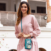 Woman in a pink outfit smiling and holding a small blue floral Anuschka genuine leather Crossbody Phone Case - 1173 with RFID card holders.