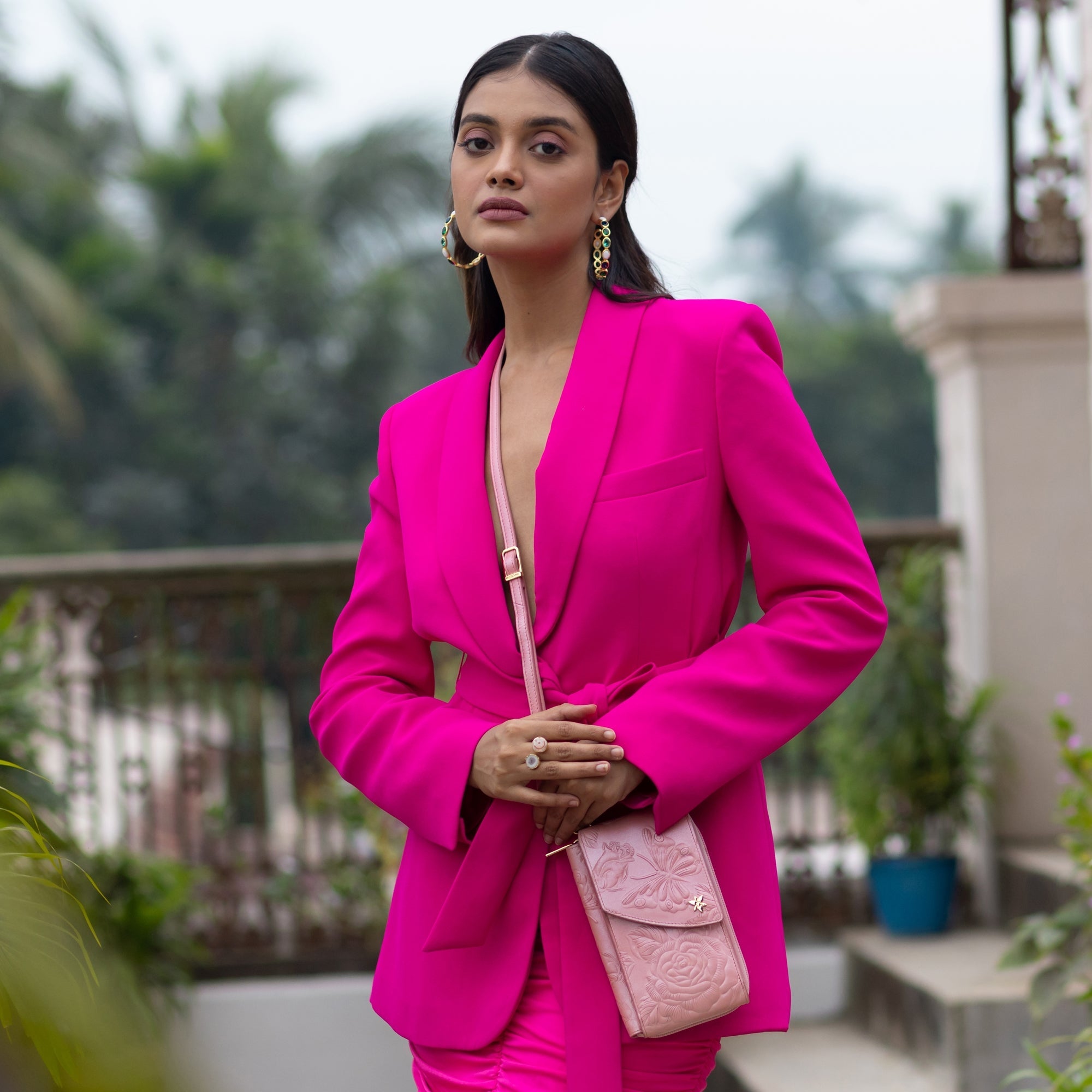 A woman in a bright pink blazer and matching pants poses outdoors, holding an Anuschka Crossbody Phone Case - 1173. She has long dark hair and wears large hoop earrings. The background includes plants and architectural elements.