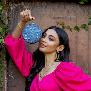 A woman in a pink outfit holding up an Anuschka Round Coin Purse - 1175 with hand-painted designs on it against a rustic backdrop.