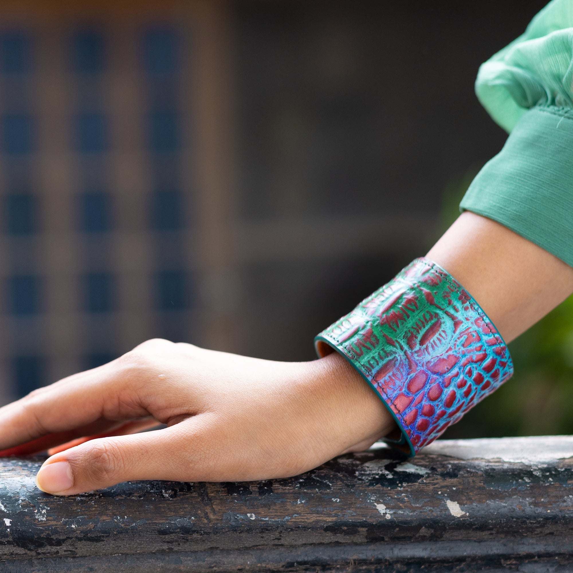 Close-up of a person's wrist wearing an Anuschka Painted Leather Cuff - 1176. The bracelet features a mix of red, purple, and green colors. The person is also wearing a green sleeve.