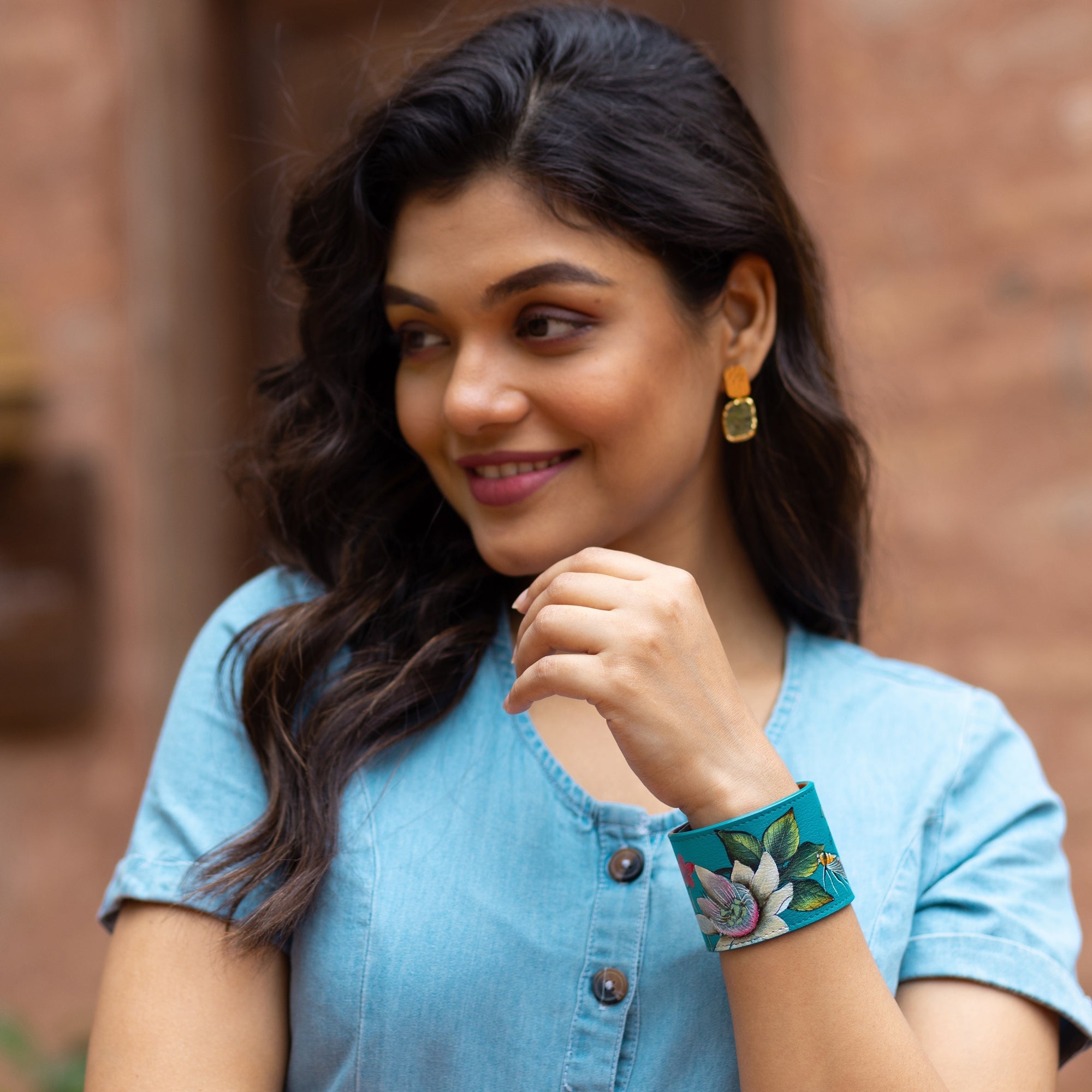 A woman with wavy hair wearing a blue button-up top and gold earrings smiles while looking off-camera, showcasing an Anuschka Painted Leather Cuff - 1176 with floral embroidery.