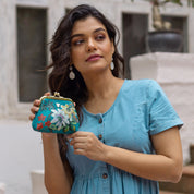 A woman in a blue dress holds an Anuschka Clasp Pouch With Key Fobs - 1177 outdoors, standing in front of a white building and greenery.