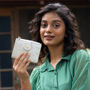 A woman with wavy hair in a green blouse holds up an Anuschka Two Fold Organizer Wallet - 1178 made of genuine leather, featuring embossed floral patterns and a decorative clasp.