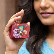 A person holding the Anuschka Airpod Pro Case - 1179, a small red leather case adorned with a colorful butterfly and flower design, made from genuine leather.