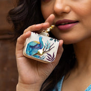 A person holds a decorative white genuine leather Anuschka Airpod Pro Case - 1179 with a peacock design near their mouth, showing only the lower half of their face and hand.