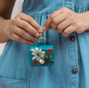 A person in a blue chambray shirt dress is holding an Anuschka Airpod Pro Case - 1179, a small turquoise floral case with a snap closure, premium hardware, and a gold clip.