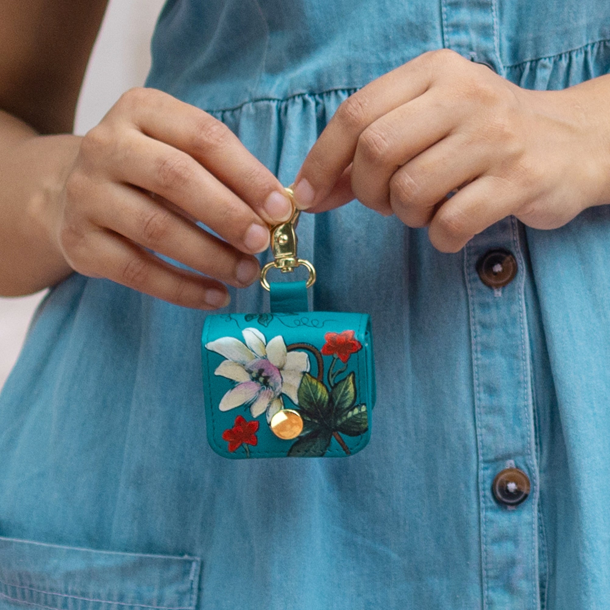 A person in a blue chambray shirt dress is holding an Anuschka Airpod Pro Case - 1179, a small turquoise floral case with a snap closure, premium hardware, and a gold clip.