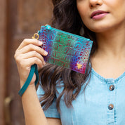 Person holding a small, colorful, patterned Anuschka Card Holder with Wristlet - 1180 made of genuine leather with a teal strap, wearing a light blue, button-down shirt. Only the lower part of the face is visible.