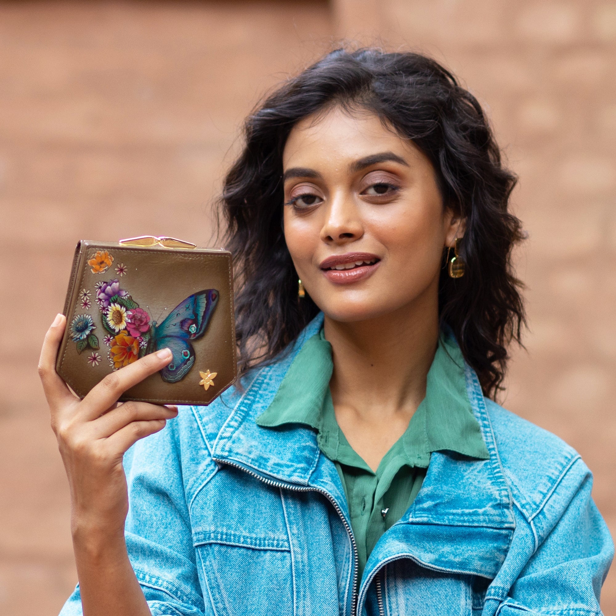 A person with shoulder-length curly hair holds up an Anuschka Two Fold French Wallet - 1181, beautifully hand-painted with a butterfly and floral design. Wearing a green shirt and denim jacket, they smile at the camera as they showcase the genuine leather wallet, which features intricate detailing that adds to its charm.