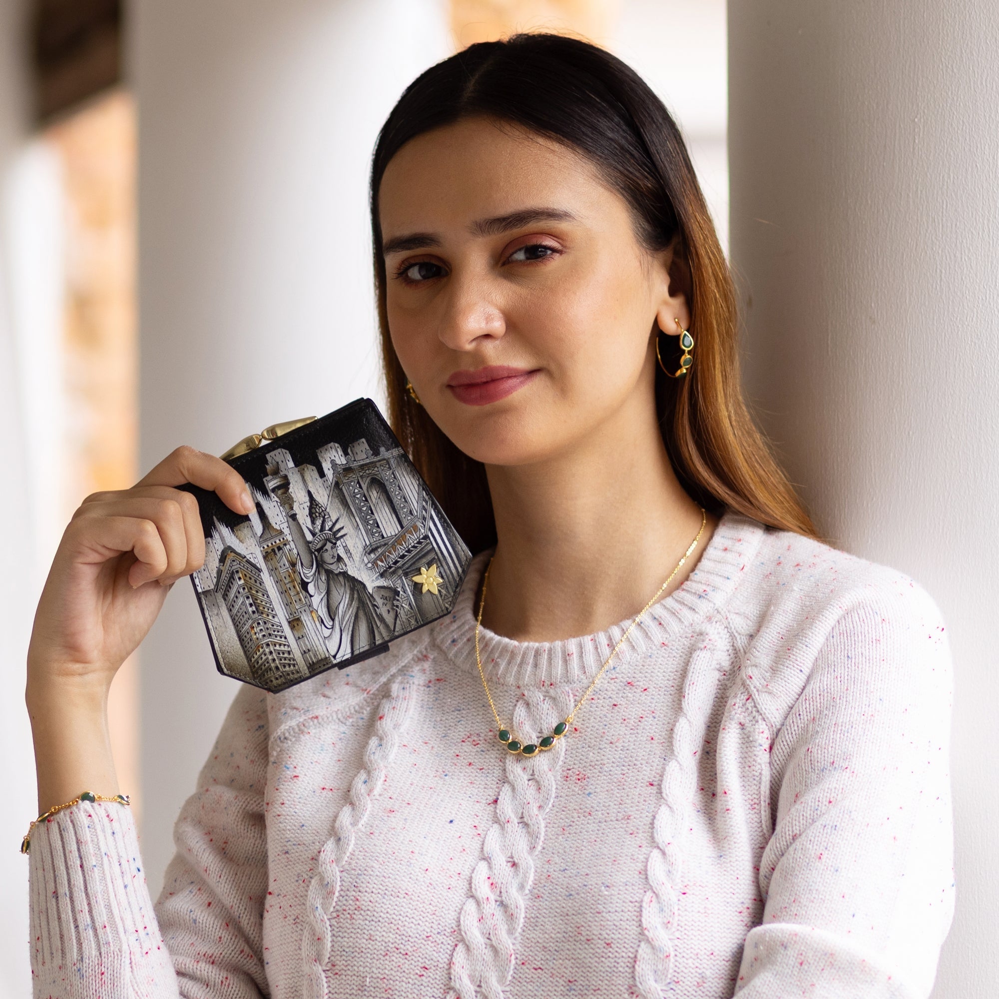 A woman wearing a light-colored sweater and jewelry holds an Anuschka Two Fold French Wallet - 1181, featuring hand-painted original artwork of the Statue of Liberty and buildings. The wallet, adorned with a French clasp, complements her elegant style as she stands against a white column.