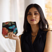 A woman in a black sleeveless top holds an Anuschka Two Fold French Wallet - 1181, adorned with hand-painted floral and butterfly designs, standing in front of a light-colored background.