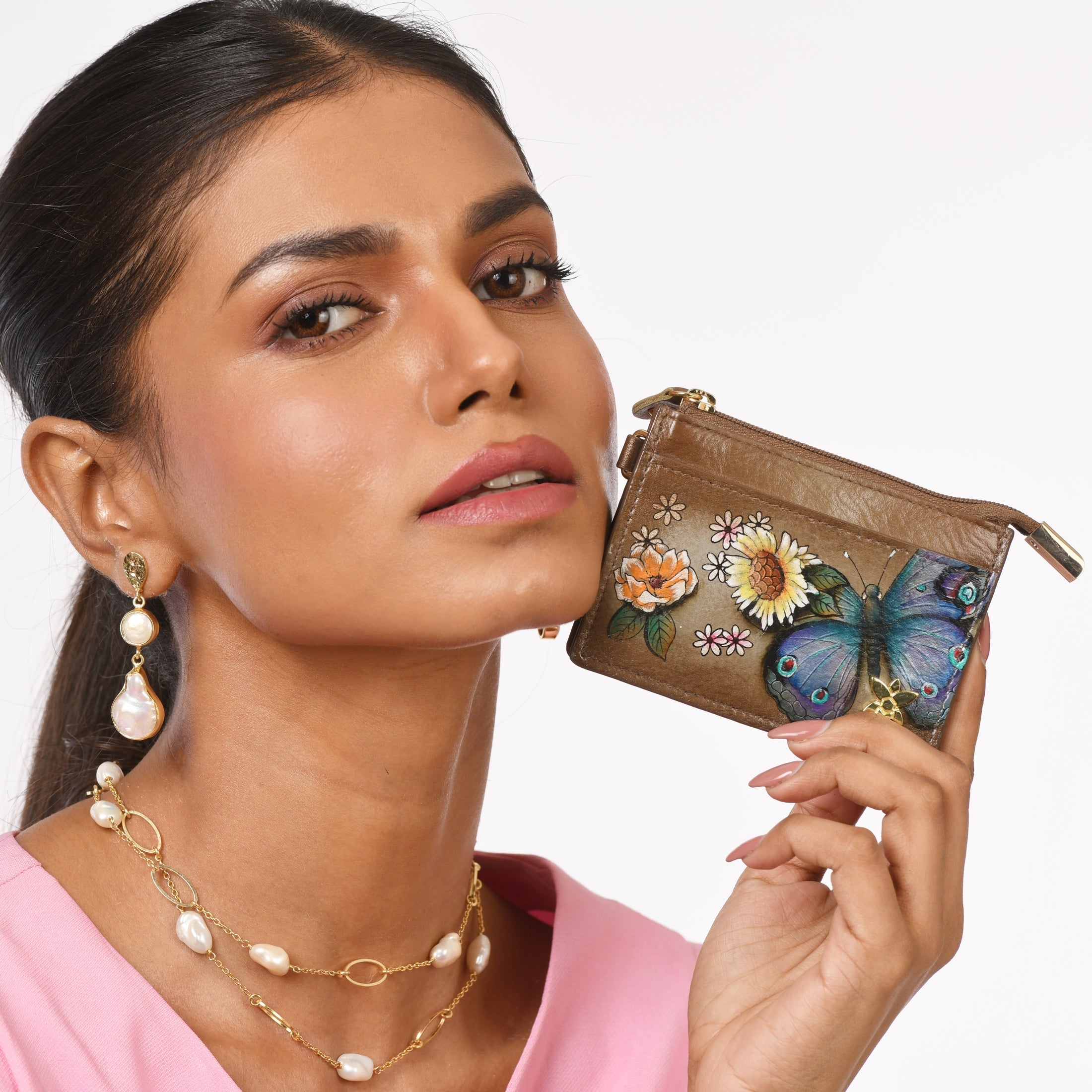 A woman in a pink top is holding Anuschka's CARD CASE - 1183, a small, hand-painted brown floral and butterfly-printed genuine leather purse. She is wearing pearl earrings and a matching necklace. This stylish and RFID-protected case adds both elegance and security to her ensemble.