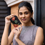 An individual softly smiles while holding up an Anuschka CARD CASE - 1183, featuring a hand-painted floral design, and is dressed in a sleeveless gray top.