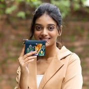 A woman holding the Anuschka CARD CASE - 1183, a small, colorful wallet with a botanical design. Hand-painted and RFID protected, this genuine leather accessory adds an elegant touch. She is standing outdoors in front of a blurred natural background wearing a tan blazer over a white top.