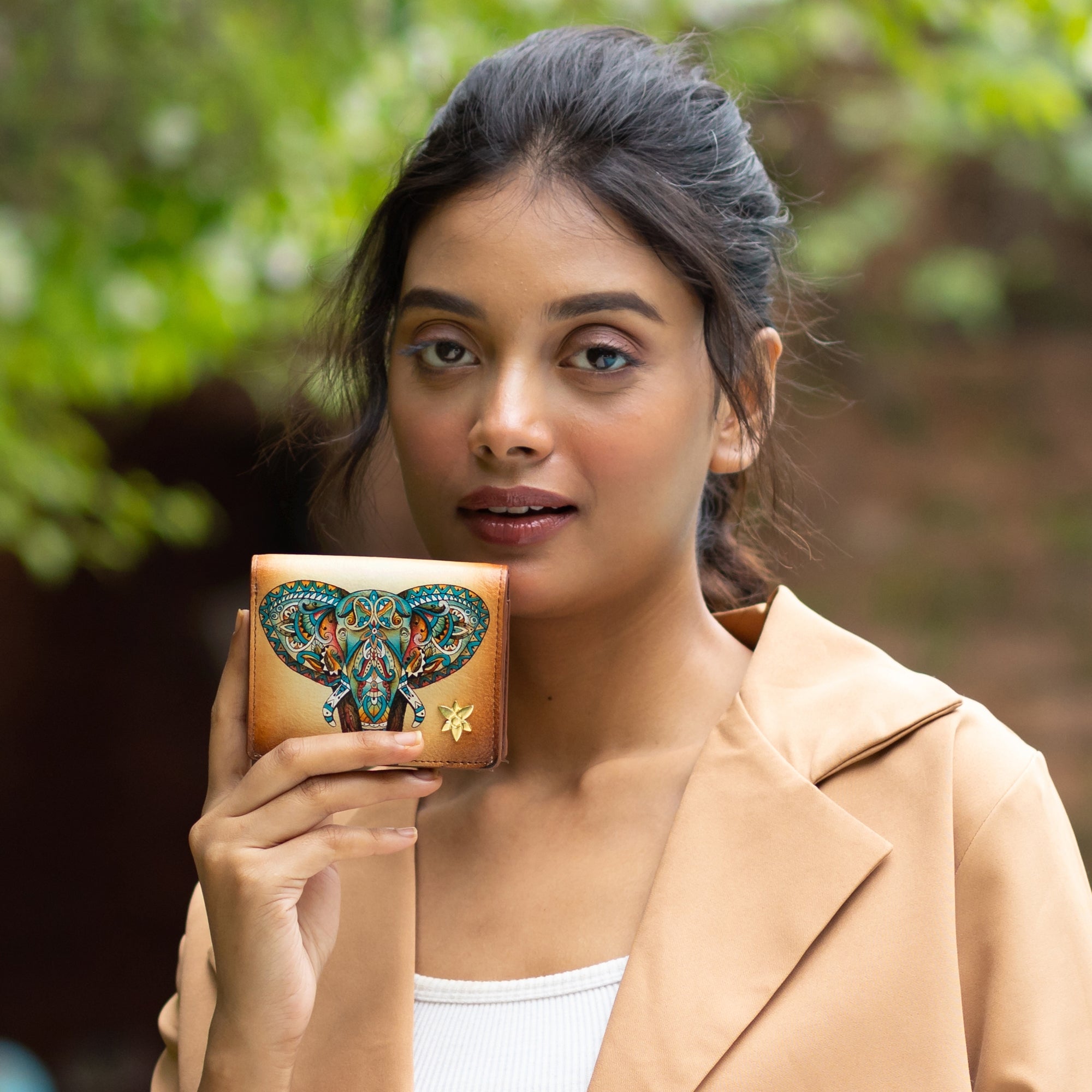 A woman holds up the Anuschka Card Organizer Wallet - 1184, showcasing its genuine leather surface adorned with a vibrant, hand-painted elephant design. She is dressed in a tan coat and a white top, standing outdoors with lush greenery as her backdrop.