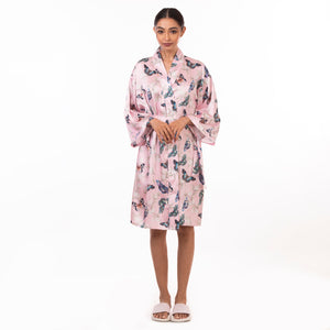 A person practicing self-care, standing against a white background, wearing a pink Anuschka 3343 robe with a bird pattern and white sandals.