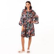 A woman models a brightly patterned silk Anuschka robe with wide sleeves, paired with white sandals, against a white background.