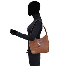 Load image into Gallery viewer, A person standing in profile with a tan Anuschka Classic Hobo With Side Pockets - 382 handbag featuring butterfly designs.
