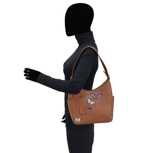 A person standing in profile with a tan Anuschka Classic Hobo With Side Pockets - 382 handbag featuring butterfly designs.