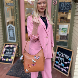 Woman posing with a brown Anuschka Classic Hobo With Side Pockets - 382 adorned with butterfly motifs, wearing a pink suit outside a shop.