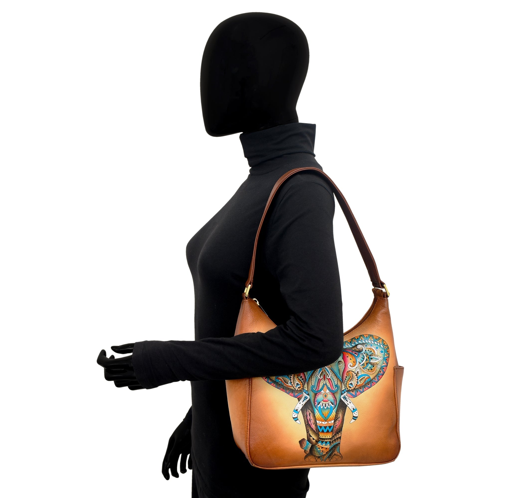 A mannequin in a black turtleneck and gloves is holding a brown Anuschka Classic Hobo With Side Pockets - 382 handbag featuring a colorful, intricate hand-painted elephant design.