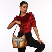 A woman in a red blouse and black pants holds the Anuschka Classic Hobo With Side Pockets - 382, a genuine leather handbag adorned with a colorful, hand-painted elephant design.