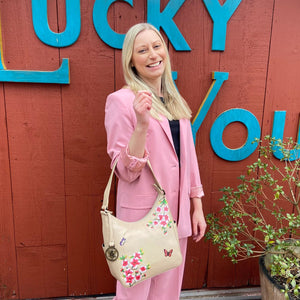 A woman in a pink suit smiling in front of a wall with the words "lucky you" written on it, holding an Anuschka Classic Hobo With Side Pockets - 382 handbag.