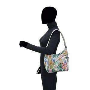 A mannequin wearing a black turtleneck and pants carries an Anuschka Classic Hobo With Side Pockets - 382 handbag with a genuine leather shoulder strap on its shoulder.
