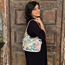 Load image into Gallery viewer, Woman smiling and posing with an Anuschka Classic Hobo With Side Pockets - 382 handbag, featuring a shoulder strap, in front of a rustic wooden door.
