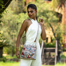 Load image into Gallery viewer, A woman in a stylish white outfit poses with an Anuschka Medium Crossbody With Double Zip Pockets - 447 hand-painted, floral patterned leather handbag.
