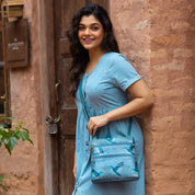 A woman in a blue dress smiling and posing with an Anuschka Medium Crossbody With Double Zip Pockets - 447 against a rustic wall.