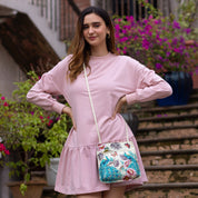 Woman in a pink dress with an Anuschka genuine leather Slim Crossbody With Front Zip - 452 standing confidently on stairs with blooming flowers in the background.