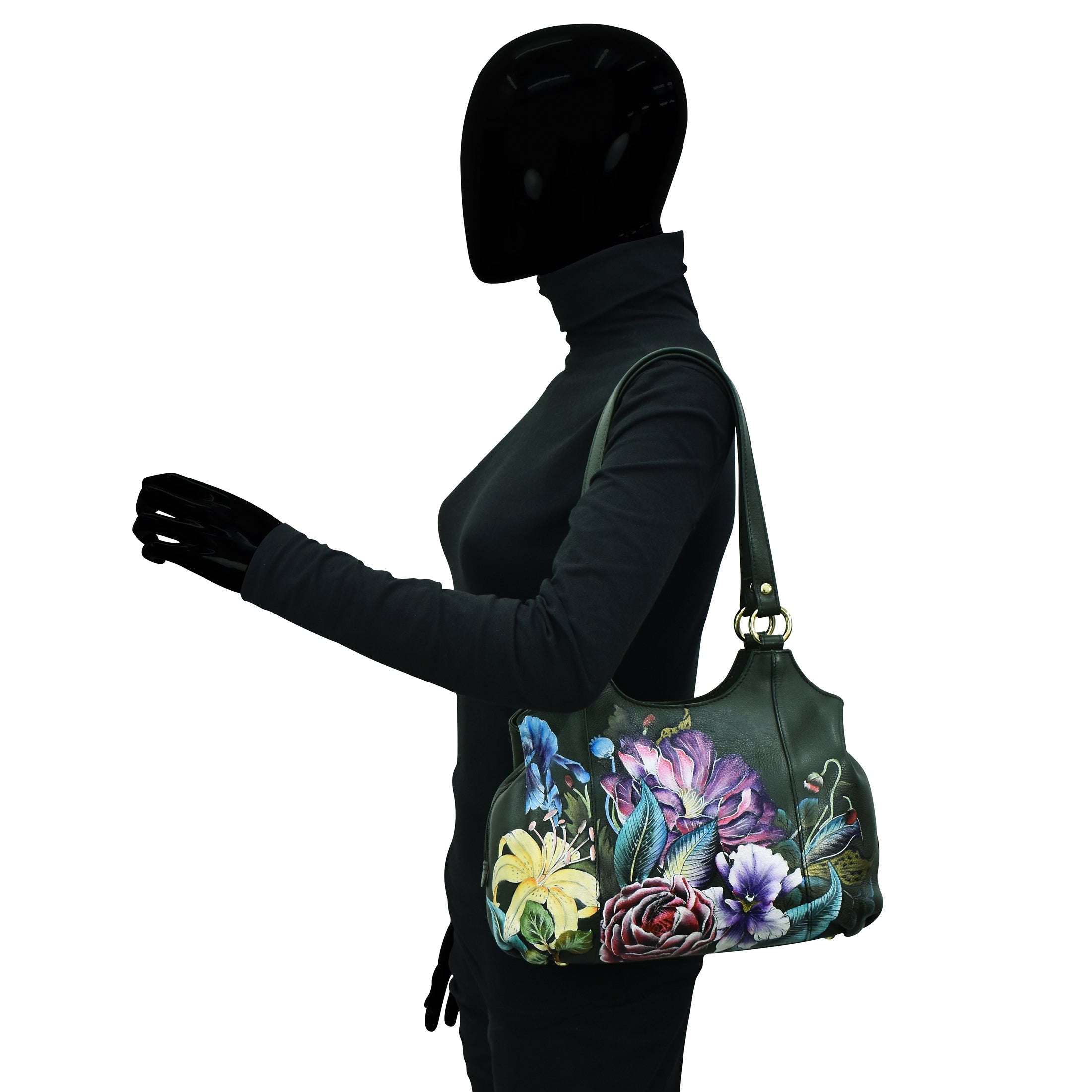 A mannequin with a smooth black face and glossy black hands models a dark floral handbag on its right shoulder while wearing a long-sleeve black turtleneck. The Triple Compartment Satchel - 469 by Anuschka adds an elegant touch to the ensemble.