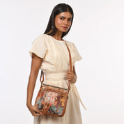 A woman wearing a light-colored dress holds an Anuschka Expandable Travel Crossbody - 550, showcasing genuine leather with colorful, ancient Egyptian-themed hand-painted artwork.