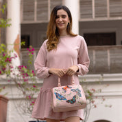 A woman in a pink dress holding an Anuschka Zip Around Classic Satchel - 625 with butterfly motifs stands in front of a building with flowers.