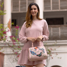 Load image into Gallery viewer, A woman in a pink dress holding an Anuschka Zip Around Classic Satchel - 625 with butterfly motifs stands in front of a building with flowers.
