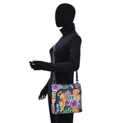 A mannequin dressed in black showcases the Anuschka Crossbody With Front Zip Organizer - 651, a hand-painted leather bag featuring a vibrant floral and leopard print design, complete with an adjustable crossbody strap.