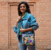 A woman in a denim jacket and jeans smiles while standing outdoors, holding an Anuschka Crossbody With Front Zip Organizer - 651 over her shoulder. She stands in front of a brick wall, showcasing the colorful floral design on her hand-painted leather crossbody with an adjustable strap.