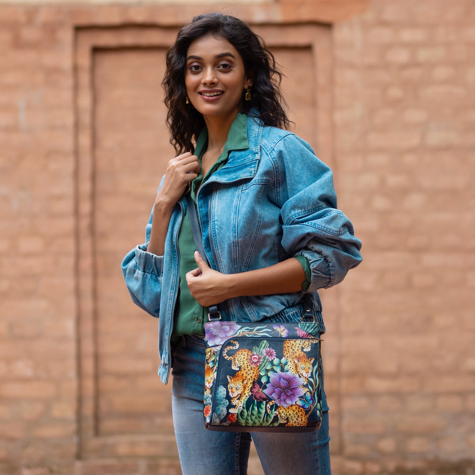 A woman in a denim jacket and jeans smiles while standing outdoors, holding an Anuschka Crossbody With Front Zip Organizer - 651 over her shoulder. She stands in front of a brick wall, showcasing the colorful floral design on her hand-painted leather crossbody with an adjustable strap.